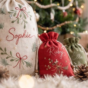 May include: Three drawstring gift bags in beige, red, and olive green linen. The beige bag features the name Sophie, the red bag features the name Ruby, and the green bag features the name Ella, all embroidered with mistletoe and bow motifs.