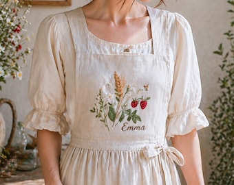 May include: A cream-colored apron with embroidered wheat, strawberries, and the name "Emma". The apron has a square neckline, puff sleeves, and pockets. A woman is holding a basket of flowers.