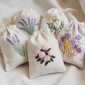以下が含まれることがあります： 刺繍された花の模様が施された、クリーム色の巾着袋が5つ。ラベンダー、デイジー、ピンクのバラ、その他のカラフルな花が描かれています。これらの袋は天然素材で作られており、白い表面に配置されています。