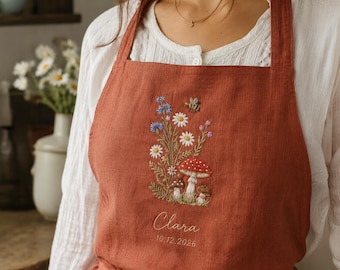 May include: A rust-colored apron featuring an embroidered design of wildflowers, mushrooms, and a bee. Below the embroidery, the name Clara and the date 10.12.2026 are stitched in white thread. The apron has thick shoulder straps and a textured fabric.
