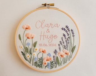 May include: Embroidered hoop art featuring the names "Clara & Hugo" and the date "18.06.2024" in pink thread. The design includes embroidered flowers in peach, blue, and white, with lavender accents. The hoop is made of wood.