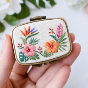 Puede incluir: Una pequeña pastillero rectangular de metal con acabado en latón antiguo, con una tapa de tela color crema bordada con un vibrante diseño floral tropical que incluye flores de hibisco rosas y naranjas, hojas de monstera y ave del paraíso.