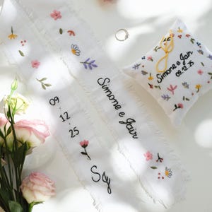 以下が含まれることがあります： 刺繍された花の模様と「Simone e Jair」の名前と日付「09.12.25」が施された白いリングピローとリボン。銀色の指輪がリボンに置かれています。背景にはピンクと白の花があります。