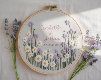 May include: Embroidered floral hoop art featuring the names "Isabelle & Johan" and the date 17.07.2025 in pink thread. The design includes white daisies and purple flowers with green stems, set against a light gray fabric. A wooden hoop frames the artwork.