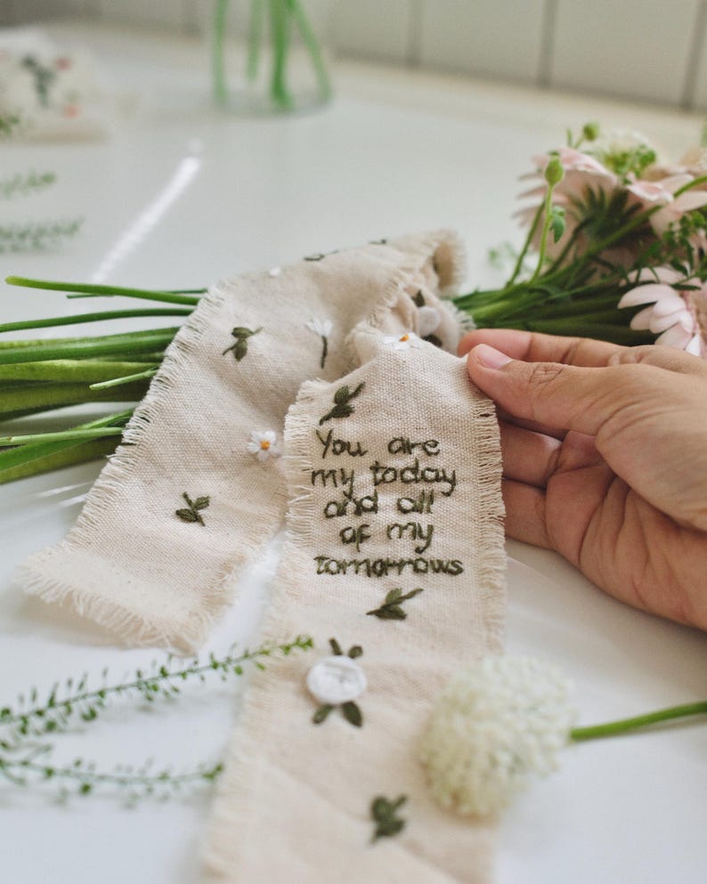 以下が含まれることがあります： クリーム色の生地タグに「You are my today and all of my tomorrows」と刺繍されています。タグには小さな刺繍の花と葉が施されています。背景にはピンクの花と緑のブーケがあります。