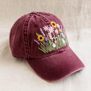 May include: A burgundy baseball cap with embroidered flowers in shades of pink, yellow, green, and purple.