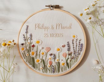 May include: Embroidered hoop art featuring the names "Philipp & Mariek" and the date "25.10.2025". The design includes embroidered flowers in white, yellow, pink, and purple, set against a cream-colored fabric. The hoop is made of wood.