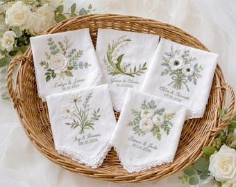 May include: Five white fabric handkerchiefs with lace edges, each featuring unique floral embroidery and personalized names and dates including Evelyn & James 05.16.2026, Grace & Oliver 06.20.2026, Chloe & Bennett 07.18.2026, Ivy & Rowan 08.29.2026, and Sienna & Jude 10.04.2026.