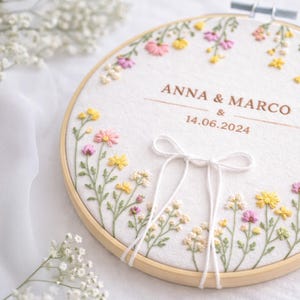 Może przedstawiać: Haft na tamborku z imionami "ANNA & MARCO" i datą "14.06.2024". Biała tkanina ozdobiona jest kolorowymi haftowanymi kwiatami w kolorze różowym, żółtym i białym, umieszczonymi w drewnianym tamborku. Na dole zawiązana jest biała kokarda.