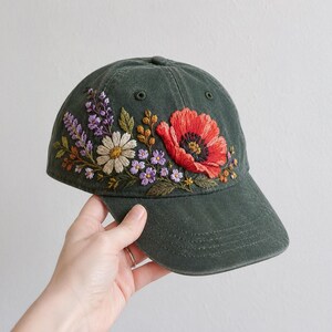May include: A dark green baseball cap featuring detailed embroidery of a large red poppy, a white daisy, purple lavender sprigs, and small blue flowers with green leaves. The floral design is stitched across the front panel of the cap.