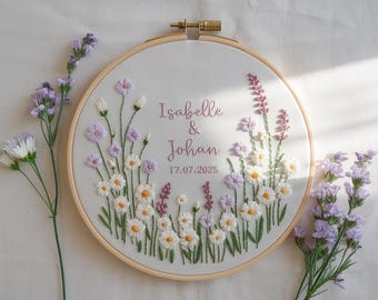 May include: Embroidered floral hoop art featuring white and purple flowers, green stems, and the names "Isabelle & Johan" with the date "17.07.2025" in a cursive font. The embroidery is set in a wooden hoop.