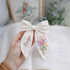 Può includere: Un fiocco di tessuto color crema con il nome "Evelyn" stampato su un lato e un ricamo floreale sull'altro. Il ricamo presenta fiori rosa, blu e gialli. Il fiocco è tenuto aperto contro un libro.