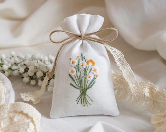 May include: A small, white linen drawstring bag with an embroidered bouquet of daisies. The daisies have white petals and yellow centers, with green stems and leaves. The bag is tied with a natural twine and lace.