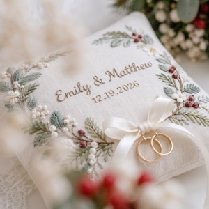 May include: A square off-white ring pillow featuring an embroidered wreath of green leaves, white flowers, and red berries. The center is embroidered with the text Emily & Matthew and 12.19.2026. A white satin ribbon holds two gold wedding bands.