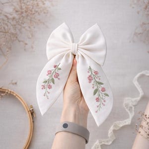 Può includere: Un grande fiocco di tessuto bianco sporco con fiori rosa e foglie verdi ricamate. Il fiocco è tenuto in alto, mostrando il ricamo floreale sulle estremità lunghe e fluenti. È un accessorio per capelli.