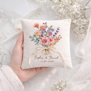 Puede incluir: Un cojín de anillos cuadrado, blanquecino, con un ramo floral bordado en tonos rosa, naranja, morado y azul. Dos anillos de oro están atados con una cinta color crema. Los nombres "Sophia & Daniel" y la fecha "12.09.2026" están bordados.
