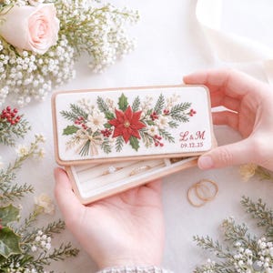 May include: A rectangular wooden ring box with a white lid featuring embroidered poinsettias, holly leaves, and red berries. The lid is personalized with the red embroidered initials L & M and the date 09.12.25. The interior has white velvet slots.