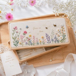 Op de afbeelding: Een houten sieradendoos met een crèmekleurig deksel met geborduurde bloemen, bijen en de tekst "Grow in love". De doos is open en onthult een compartiment met ringen en een ketting. De doos staat op een houten dienblad.