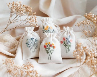 May include: Four small, off-white drawstring bags with embroidered floral designs. The bags feature various colorful flower arrangements in red, yellow, blue, and pink. The bags are arranged on a soft, white fabric, with dried flowers in the background.