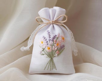 May include: A white linen sachet with a bouquet of embroidered flowers. The bouquet includes lavender, daisies, and other wildflowers in shades of pink, purple, and yellow. The sachet is tied with a twine bow and has lace trim.
