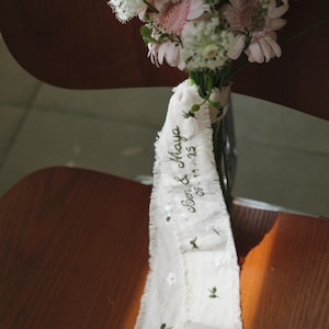 以下が含まれることがあります： ピンクと白の花束と装飾リボン。リボンはクリーム色で、縁がほつれており、「Ben & Maya 09.11.25」の刺繍と小さな花のディテールが施されています。花束は透明なガラスの花瓶に入っています。