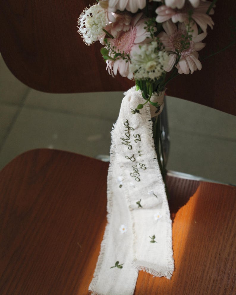 以下が含まれることがあります： ピンクと白の花束と装飾リボン。リボンはクリーム色で、縁がほつれており、「Ben & Maya 09.11.25」の刺繍と小さな花のディテールが施されています。花束は透明なガラスの花瓶に入っています。