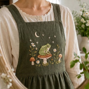 May include: An olive green apron with embroidered details. The design features a frog sitting on a mushroom, with flowers, stars, and a crescent moon. The apron has a square neckline and wide straps.