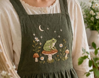 May include: An olive green apron with embroidered details. The design features a frog sitting on a mushroom, with flowers, stars, and a crescent moon. The apron has a square neckline and wide straps.