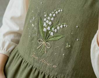 May include: Olive green apron with a floral embroidery of white lily of the valley flowers and green leaves. The apron has a ruffled hem and the text "Juin Atelier" embroidered in a cursive font. Small white beads are scattered around the floral design.