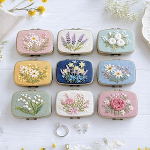 Könnte beinhalten: Neun rechteckige, bestickte Schmuckkästchen mit floralen Motiven in verschiedenen Farben wie Rosa, Gelb, Blau und Grün. Jede Schatulle zeigt einzigartige, handgestickte Blumenmuster wie Gänseblümchen, Lavendel und Rosen. Ein silberner Ring und zwei kleine Ohrstecker liegen darunter.