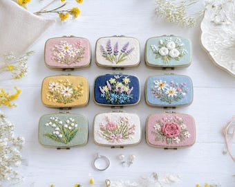 May include: Nine rectangular embroidered jewelry boxes featuring floral designs in various colors including pink, yellow, blue, and green. Each box displays unique hand-stitched flower patterns such as daisies, lavender, and roses. A silver ring and two small stud earrings are placed below.