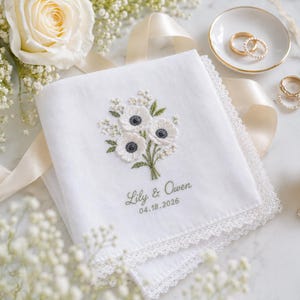 May include: A white square handkerchief with a lace border, featuring an embroidered bouquet of white anemone flowers with dark centers and green stems. Below the flowers, the text reads Lily & Owen and the date 04.18.2026 in green thread.