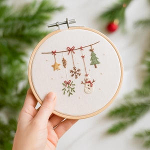 May include: Embroidered Christmas hoop art featuring festive designs. The artwork includes a gold star, pinecone, snowflake, Christmas tree, snowman, and mistletoe, all suspended from a brown thread. The embroidery is set in a wooden hoop.