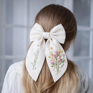 Può includere: Un grande fiocco per capelli bianco sporco con lunghe code. La coda sinistra è ricamata con il nome "Amelia" in verde. La coda destra presenta un ricamo floreale con rose rosa, accenti gialli e piccole perle.