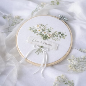 Peut inclure: Un cercle de broderie blanc avec un bouquet de fleurs blanches et de feuilles vertes. Le texte "Elise & Matteo" est brodé sur une bannière blanche. Un ruban blanc est noué sous le bouquet. Le cercle est en bois.