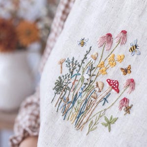Op de afbeelding: Een lichtbeige linnen stof met een gedetailleerd borduurontwerp van wilde bloemen, paddenstoelen, bijen en een vlinder. De botanische scène bevat roze zonnehoeden, gele bloesems, een rood-gekapte paddenstoel en diverse groene stengels.
