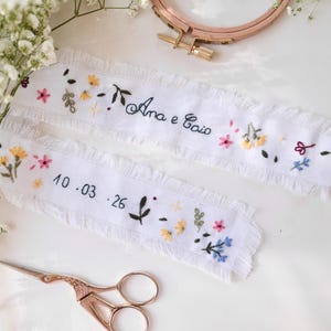 May include: Two white embroidered fabric strips with floral and leaf designs. The text "Ana e Caio" and the date "10.03.26" are embroidered on the strips. A pair of rose gold scissors and a floral arrangement are also in the image.