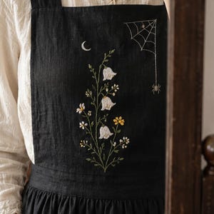 Könnte beinhalten: Eine schwarze Schürze mit einer aufwendigen Stickerei eines zentralen Blumenstraußes mit weißen glockenförmigen Blüten und gelben Blumen. Über den Blumen sind ein kleiner weißer Halbmond und ein Spinnennetz mit einer hängenden Spinne eingestickt.