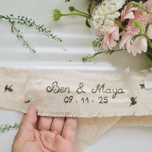 以下が含まれることがあります： クリーム色の布リボン。縁はほつれており、名前「Ben & Maya」と日付「09.11.25」が濃い緑色の糸で刺繍されています。小さな花の刺繍がリボン全体に散りばめられています。ピンクと白の花のブーケが背景にあります。