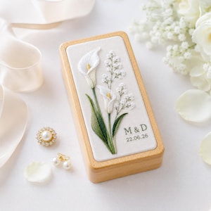 以下が含まれることがあります： 白いカラーの花と小さな白い花が刺繍された蓋を持つ長方形の木製ボックス。刺繍にはダークグリーンの糸で「M & D」の文字と「22.06.26」の日付が施されています。ボックスは滑らかな質感のライトブラウンの木製です。