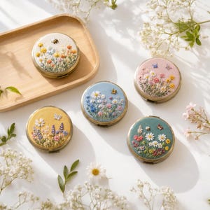 May include: Five round compact mirrors featuring intricate floral embroidery on their covers. The designs include various colorful wildflowers, butterflies, and bees stitched onto white, light blue, yellow, pink, and sage green fabric backgrounds.
