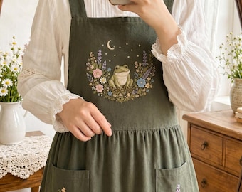 May include: An olive green apron featuring a central embroidered design of a frog surrounded by colorful flowers and a crescent moon. The apron has two front pockets, each decorated with small floral embroidery, and wide shoulder straps.