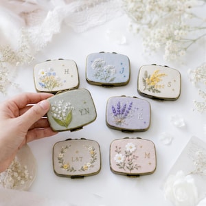 Puede incluir: Una colección de siete espejos de bolsillo rectangulares de metal con tapas de tela bordada. Cada espejo muestra diseños florales únicos en varios colores, incluyendo lavanda, blanco y amarillo, junto a iniciales bordadas como H, J, S, Y.N, K, L&T y M.