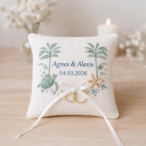 May include: A square, off-white ring pillow with embroidered palm trees, a sea turtle, and a starfish. The names "Agnes & Alexis" and the date "04.03.2026" are embroidered in blue. Two gold rings are tied with a white ribbon.
