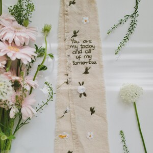 以下が含まれることがあります： 刺繍された花柄と葉柄のデザインが施されたベージュ色の布リボン。テキストは「You are my today and all of my tomorrows」です。リボンはピンク色のガーベラとその他の緑に囲まれています。