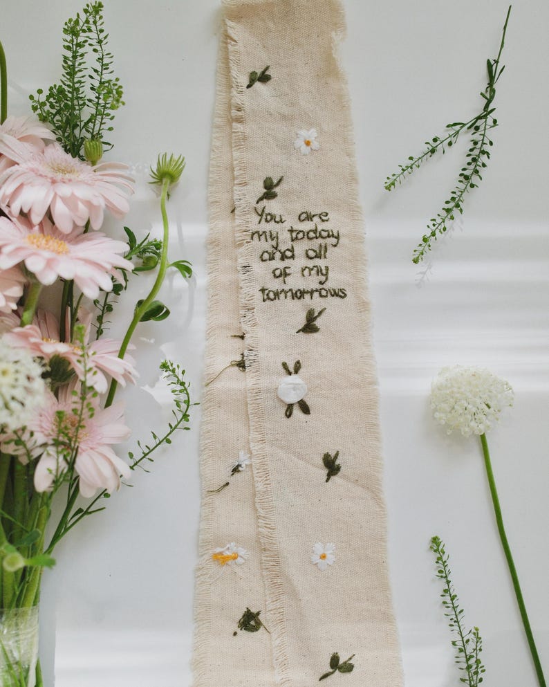 以下が含まれることがあります： 刺繍された花柄と葉柄のデザインが施されたベージュ色の布リボン。テキストは「You are my today and all of my tomorrows」です。リボンはピンク色のガーベラとその他の緑に囲まれています。