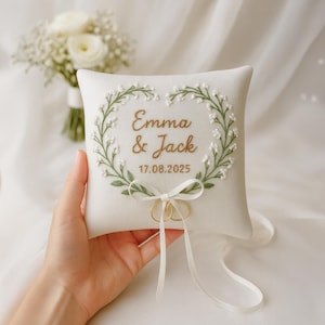 May include: A square, off-white ring pillow with embroidered floral wreath and the names "Emma & Jack" and the date "17.08.2025" in gold thread. Two gold wedding rings are attached with a white ribbon. A bouquet of white flowers is in the background.