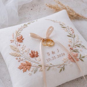 May include: A square white ring bearer pillow featuring an embroidered wreath of autumn leaves in shades of orange, brown, and green. The center includes the embroidered text S & J and 10.25.2026, with two gold rings tied by a light beige ribbon.