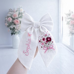 Può includere: Un fiocco per capelli in tessuto bianco con dettagli ricamati. Un lato presenta il nome "Sophia" in corsivo rosa con una farfalla. L'altro lato mostra un bouquet floreale nei toni del rosa e del bordeaux, legato con un fiocco rosa. Il fiocco è tenuto.