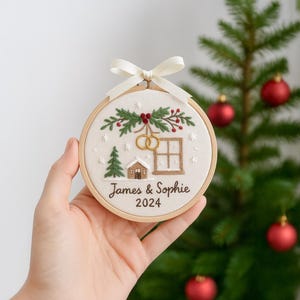 May include: Embroidered Christmas ornament in a wooden hoop, featuring a winter scene with a house, tree, and wedding rings. The text reads "James & Sophie 2024". A white ribbon is tied at the top for hanging.